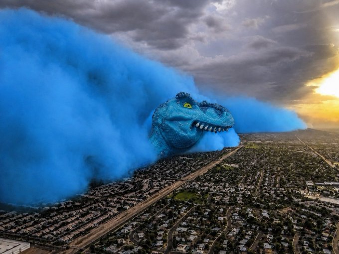 A surreal digital artwork depicting a massive blue dinosaur head with glowing yellow eyes emerging from blue smoke clouds over a sprawling suburban landscape at sunset.