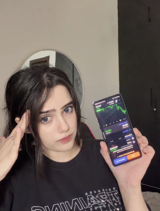 A young woman with long dark hair wearing a black t-shirt holds up a smartphone displaying a trading chart, posing for the camera indoors.