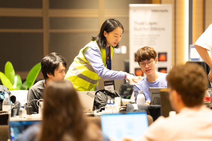 A busy conference or workshop scene where a woman in a yellow safety vest leans over to assist two young attendees working on laptops at a table. A partially visible presentation board stands in the background.