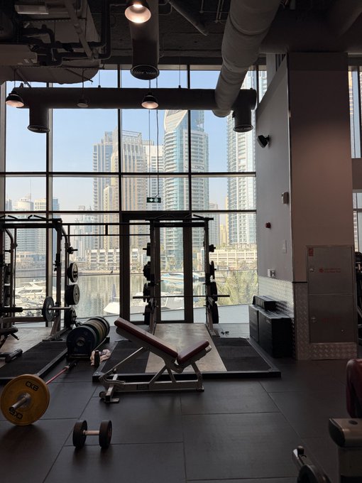 An empty gym with weight benches, dumbbells, and a squat rack, featuring large floor-to-ceiling windows overlooking a city skyline with high-rise buildings.