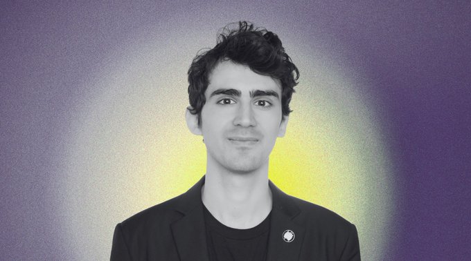 Black and white portrait photo of a young man with dark curly hair wearing a blazer over a dark shirt with a small pin, set against a purple background with a soft yellow glow behind him.