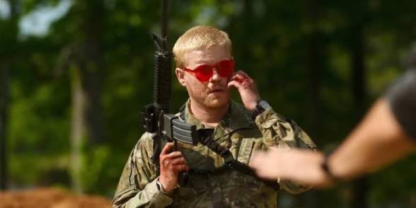 A young man with red hair and red-tinted sunglasses wearing military camouflage gear and holding an assault rifle, appearing to be at an outdoor shooting range.