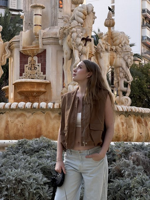 Photo of a young woman with long blonde hair wearing a brown sleeveless vest and light jeans, standing in front of an ornate baroque stone fountain with sculptural details.