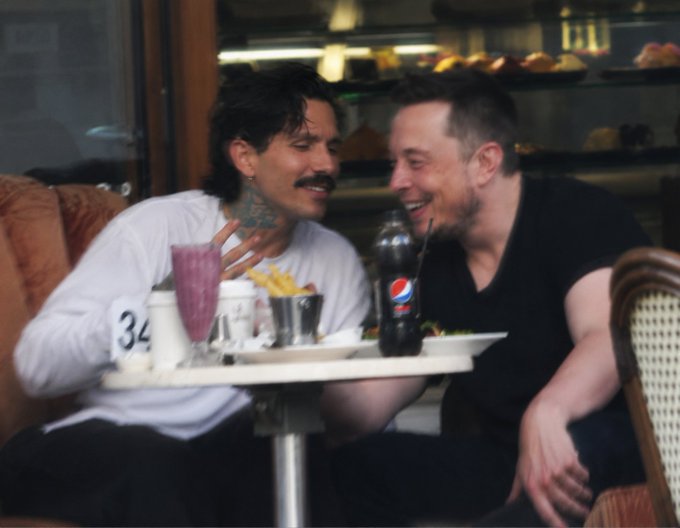 Two men laughing together at a café table with fries and a Pepsi drink, one wearing a white shirt with '34' and the other in a black t-shirt.