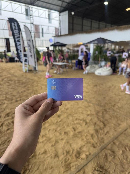 A hand holds a blue/purple gradient Visa credit card at what appears to be a beach-themed event or venue with sand and people in the background.
