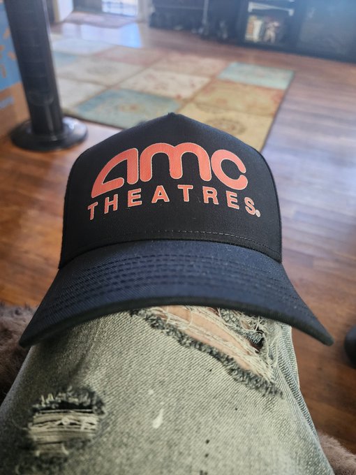 A black baseball cap with an orange embroidered "AMC THEATRES" logo rests on a person's ripped denim-clad knee, with a patterned area rug and hardwood floor visible in the background.