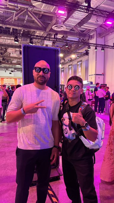 Two men wearing circular 'Meta' sunglasses pose at a convention, with one making a peace sign and the other a thumbs-up.