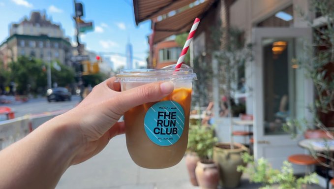The image shows a hand holding up a cup of coffee with an orange logo, possibly from "The Run Club", as indicated by the text in the photo. The background features a city street scene with buildings and cars, suggesting that this location is likely a cafe or restaurant. The caption reads "We want whatever the people who run encrypted miles at 9am have". This implies that the coffee shop offers a unique experience for runners who wish to enjoy their coffee while running in the area.