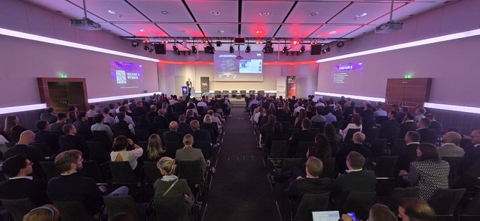 A wide shot of a large conference or seminar room with red lighting, showing a speaker presenting on stage in front of a seated audience.