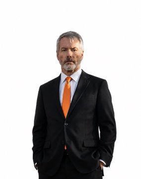 A middle-aged man with gray hair and a beard, wearing a black suit, white shirt, and orange tie, stands confidently with his hands in his pockets against a white background—appearing to be Michael Saylor, the executive referenced in the post discussing his company's $STRC preferred stock goal of returning to par.