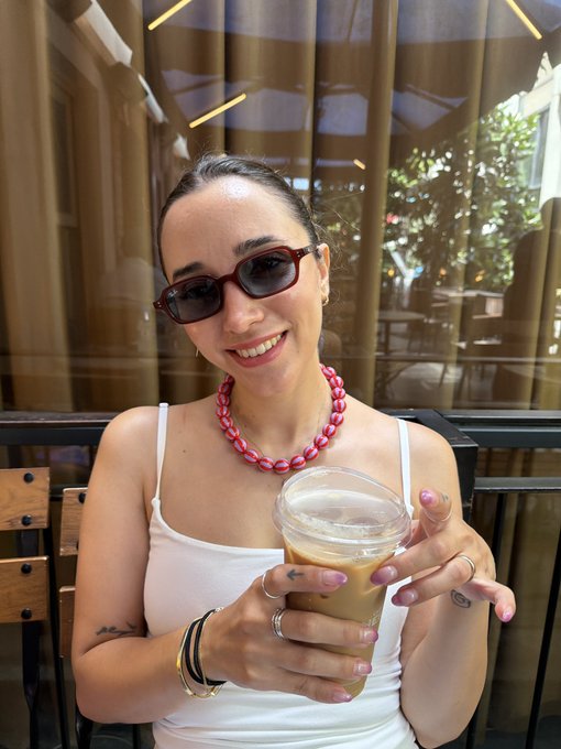 A smiling woman wearing dark sunglasses and a pink beaded necklace holds an iced coffee drink while seated at what appears to be a café.