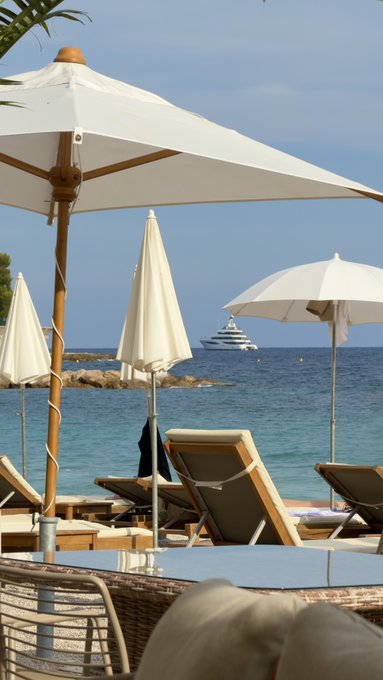 A beachside scene with white sun umbrellas, wooden lounge chairs, and a large yacht visible on the turquoise sea in the distance.