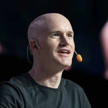 A bald man wearing a black t-shirt with a headset microphone, appearing to be giving a presentation or speech on a stage.