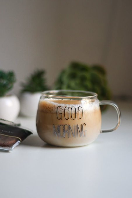 A clear glass mug filled with a frothy coffee beverage sits on a white surface, engraved with the words 'GOOD MORNING', with small potted plants blurred in the background.