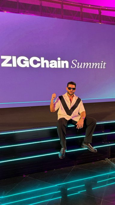 A man in a stylish white and black shirt and sunglasses poses confidently on illuminated neon steps in front of a vibrant purple backdrop displaying "ZIGChain Summit," visually reinforcing the bullish $ZIG narrative by showcasing his presence at the project's official event where the highlighted catalysts—revenue flywheel, burns, buybacks, and institutional partnerships—are taking shape.