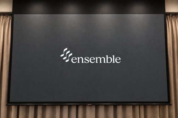 A wall-mounted display screen showing the 'ensemble' logo against a dark background, framed by tan curtains.