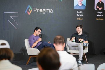 Two men sit on stage during the Pragma Lisbon event, smiling and laughing in conversation, with the audience visible in the foreground and the names "Kartik Talwar (Pragma)" and "Diogo Mónica" displayed on the screen behind them.