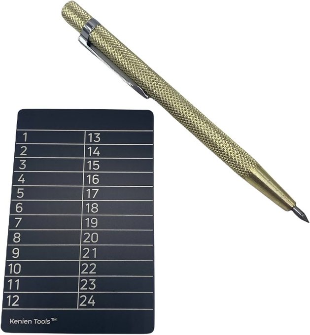 A brass knurled scribe/engraving pen rests atop a black Kenien Tools™ numbered reference card (1-24), likely used as a DIY solution for permanently marking crypto seed phrases or private keys onto metal for secure offline storage—an improvised alternative to commercial hardware wallets.