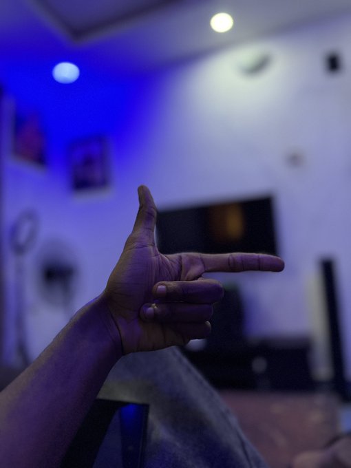 A person's hand making a finger gun gesture in a dimly lit room illuminated by blue and warm light, with a television screen visible in the background.