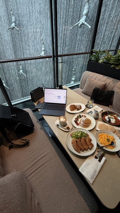 An overhead view of a breakfast spread on a table by a window with a bamboo water feature, featuring a laptop showing a chart, plates of food including eggs and meat, coffee, and bags.
