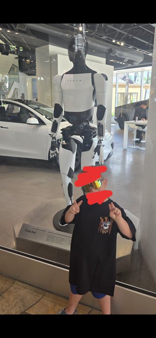 A child poses with both fingers up in front of a glass display featuring the "Tesla Bot" humanoid robot. The image relates to the post about the child wanting an Optimus robot, reflecting excitement about the Tesla humanoid robot showcased in the background.