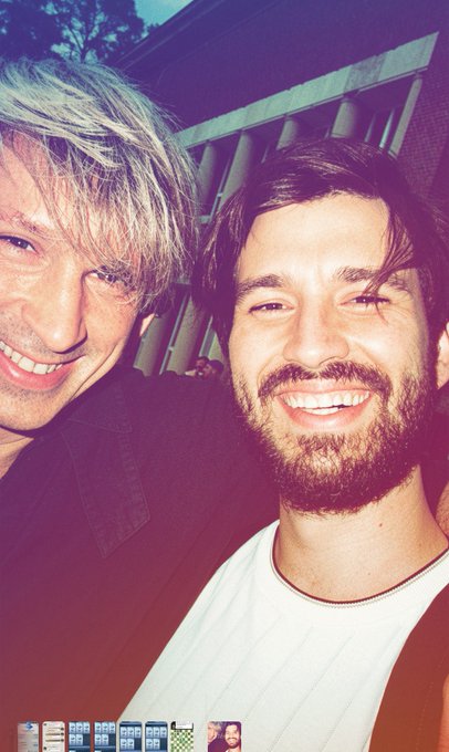 The image features two men posing together for a photo with their smiles radiating happiness. The man on the left has blonde hair and is wearing a black jacket, while the man on the right has a beard and is dressed in a white shirt. They are both smiling at the camera, creating an endearing moment captured in time.