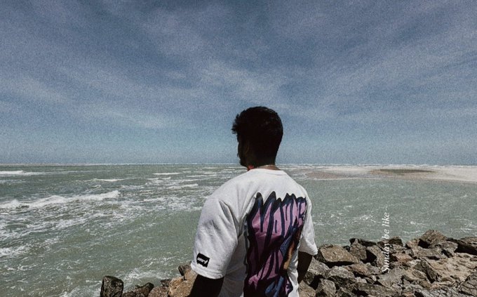 A person wearing a white graphic t-shirt stands facing a turbulent sea under a cloudy sky, viewed from behind near a rocky shore.