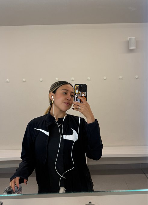 A young woman takes a mirror selfie wearing a black Nike windbreaker and white wired earphones, with a phone case featuring cartoon characters.