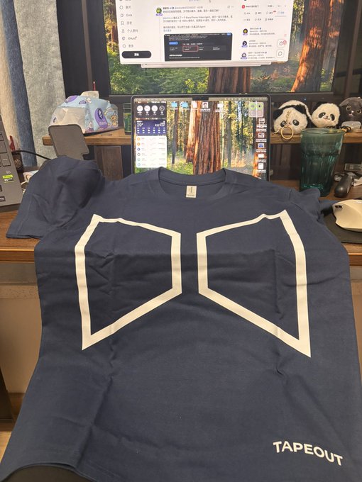 A dark navy t-shirt with a white geometric logo and 'TAPEOUT' text sits on a desk in front of a monitor, surrounded by panda plushies and a gaming setup.