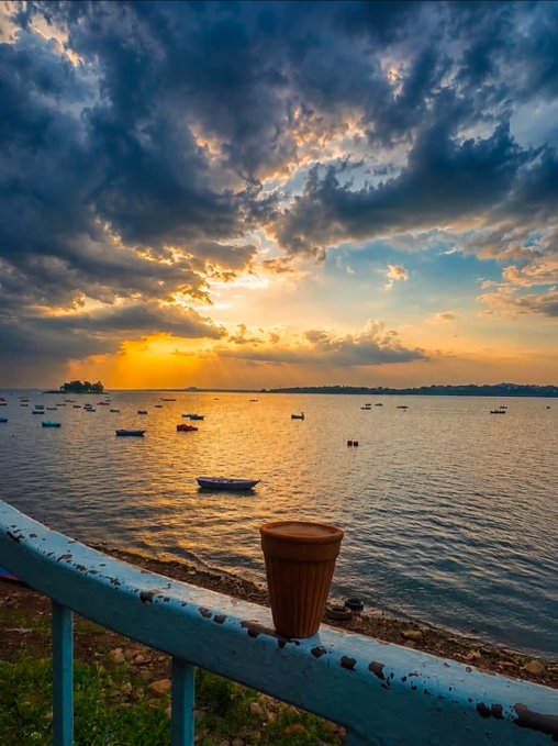 A sunset over the ocean features dramatic clouds, sun rays, small boats, and a plant pot resting on a weathered blue railing in the foreground.