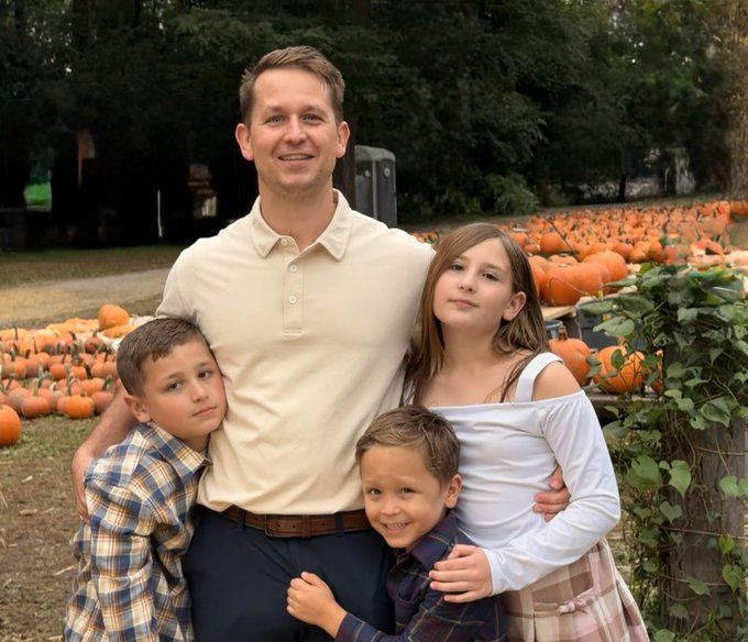 A family of four posing together at a pumpkin patch, with a father in a cream polo shirt standing with three children, surrounded by orange pumpkins and greenery.
