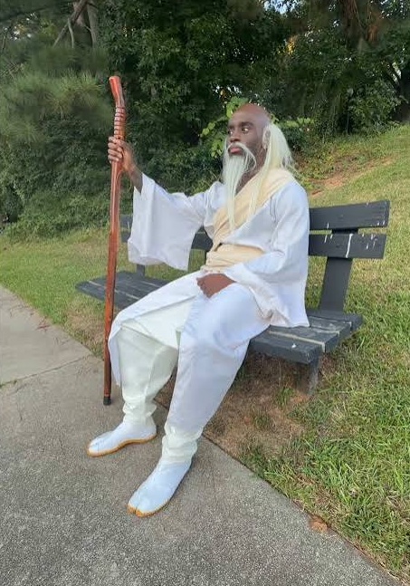A person dressed as the wizard Saruman from Lord of the Rings, wearing white robes and a long white beard, sitting on a park bench while holding a wooden staff.