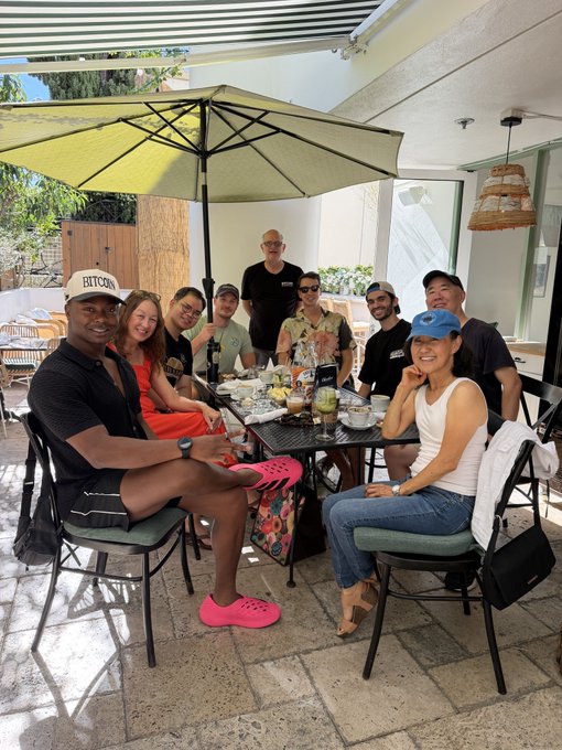 A group of about nine people gather around a table at an outdoor restaurant under a green umbrella, posing together for a photo during a meal.