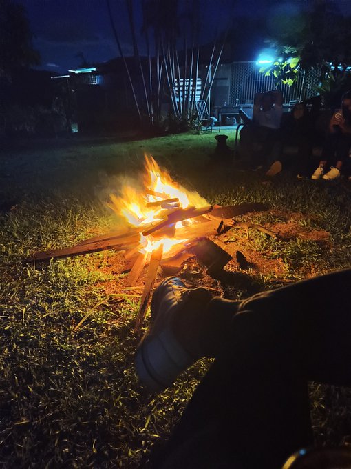 A nighttime photograph shows a campfire burning brightly in a grassy yard with people sitting nearby in the background, while a person's shoe is visible warming by the flames in the foreground.