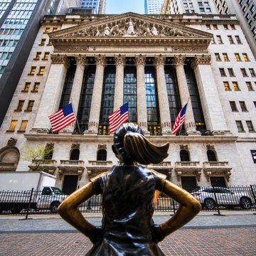A bronze "Fearless Girl" statue stands with hands on hips facing the New York Stock Exchange, whose facade displays American flags and "NEW YORK STOCK EXCHANGE" signage—symbolizing the intersection of traditional finance and the crypto exchange OKX's new push, alongside ICE, to tokenize 63 NYSE-listed stocks on-chain.