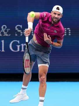 A tennis player in a maroon shirt and grey shorts serving a tennis ball on a blue court.