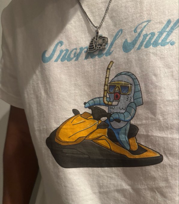 A white "Snorkel Intl." t-shirt features a cartoon fish character wearing a snorkel and goggles while riding a yellow jet ski, with a silver chain necklace draped over it. This merchandise release ties into the Snorkel Explorers IP project on $SUI, showcasing the branded apparel teased as part of building out the project's lore and identity for Year 2.