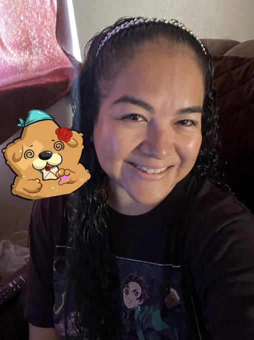 Selfie of a smiling woman with curly dark hair wearing a black headband and Demon Slayer t-shirt, with a cartoon Pomeranian sticker with a rose and party hat on the side.