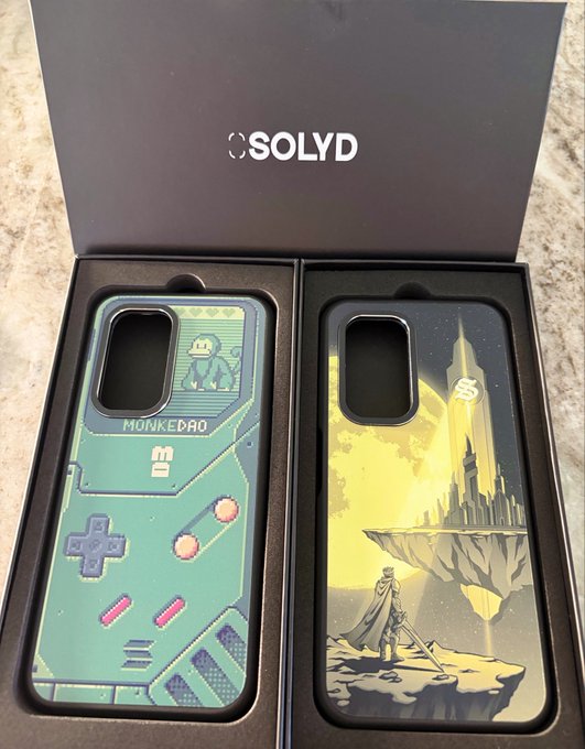 A SOLYD branded box containing two phone cases: one features a retro gaming handheld design with 'MONKEDAO' and a monkey graphic, and the other depicts a fantasy scene of a character looking at a floating castle against a yellow sky.