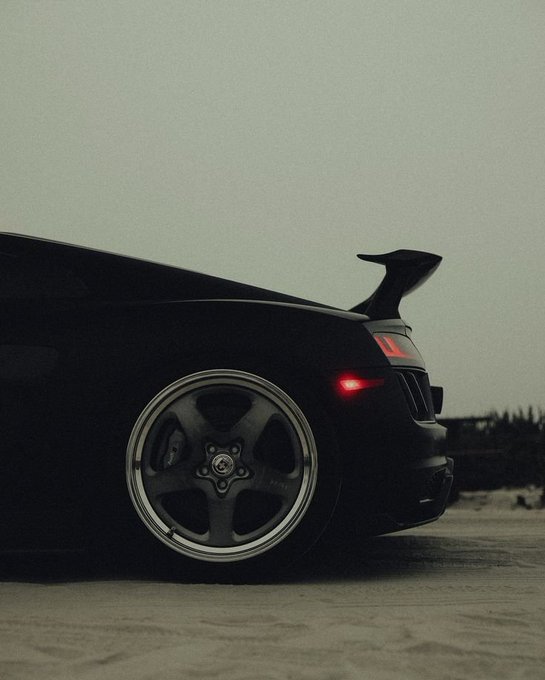 A moody, low-angle shot shows the rear of a black sports car with a prominent spoiler and aftermarket wheels parked on sand under a hazy sky.