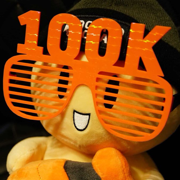A plush Bitcoin mascot wearing orange shutter shades and a beanie celebrates the milestone, with the text "100K" displayed prominently across the image to mark Bitcoin reaching $100,000.