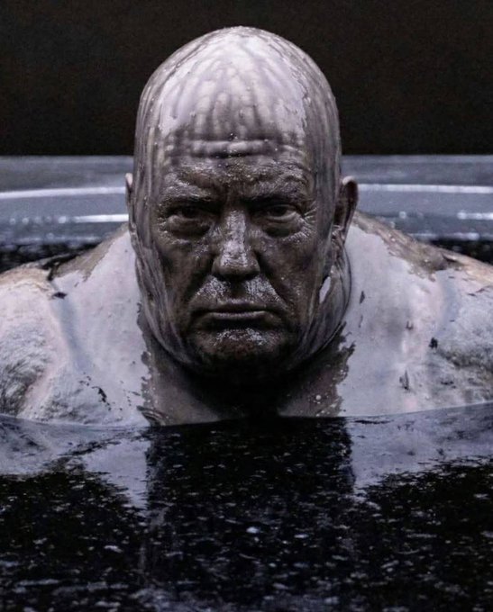 A bald, scarred figure with a grim expression emerges from dark liquid in what appears to be a promotional or cinematic still image.