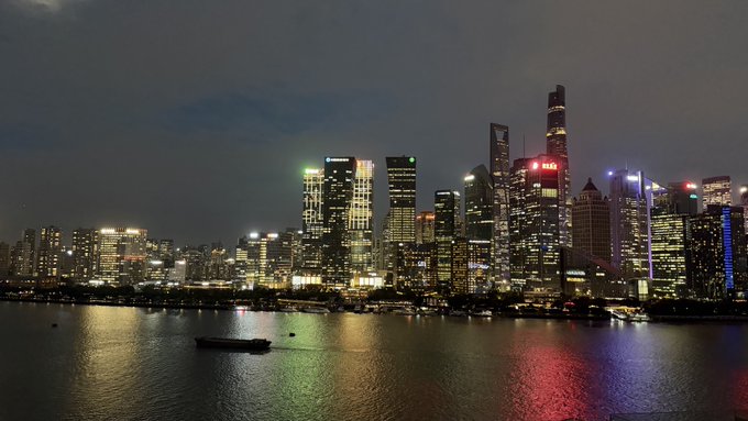 A nighttime cityscape of Shanghai's Pudong skyline featuring illuminated skyscrapers including the Shanghai Tower, with city lights reflecting on the Huangpu River below.