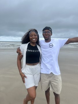A couple posing together on a beach under overcast skies; the woman wears a black 'EMBODI' crop top and checkered shorts, while the man wears a white t-shirt and khaki shorts with a black cap.