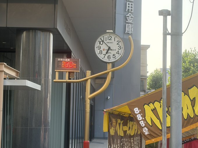 A Seiko clock mounted on a gold pole outside a Japanese building (likely a bank/credit union based on 用金庫 signage), displaying 20:03 in red digital text, with a yellow tent visible in the foreground.