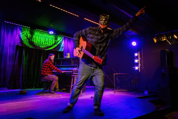 A performer wearing a fox mask plays an acoustic guitar on stage at Westward Music Festival, with a pianist in a red plaid shirt behind them under purple and blue lighting.