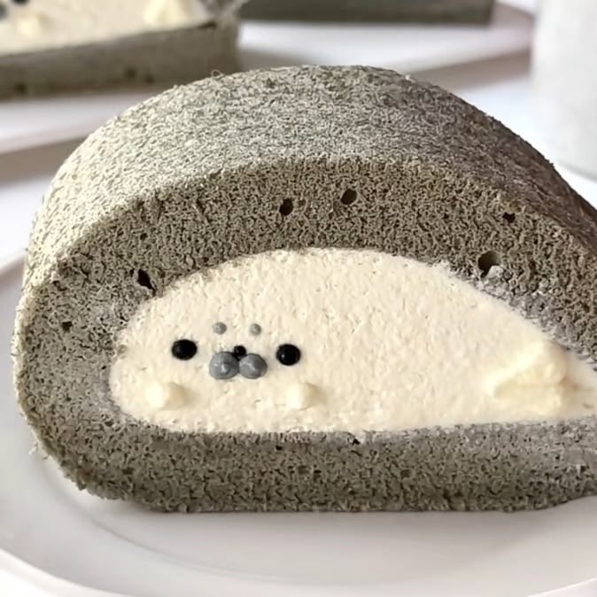 A slice of gray roll cake with white cream filling designed to look like the character Pusheen the cat, with the face created using chocolate chips and icing.