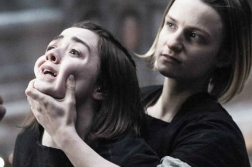 A dramatic cinematic still showing two women with dark hair in dark clothing; one woman grips the other's chin, appearing distressed or emotional in a tense moment.