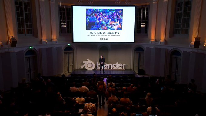 A presenter stands on stage at Blender Conference 2026 in Amsterdam, with a screen behind him displaying "THE FUTURE OF RENDERING" alongside the Render Network and Blender logos, addressing an attentive audience seated in a grand hall.