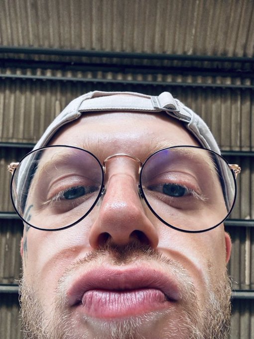 A close-up selfie of a man with round glasses and a backwards cap making a playful expression.